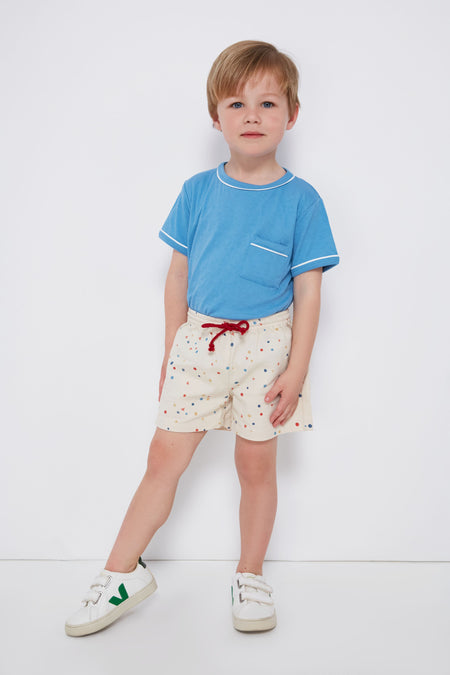 A young child wears an Oso & Me blue unisex jersey t-shirt, cream shorts with multicolor dots, and white sneakers featuring green details.