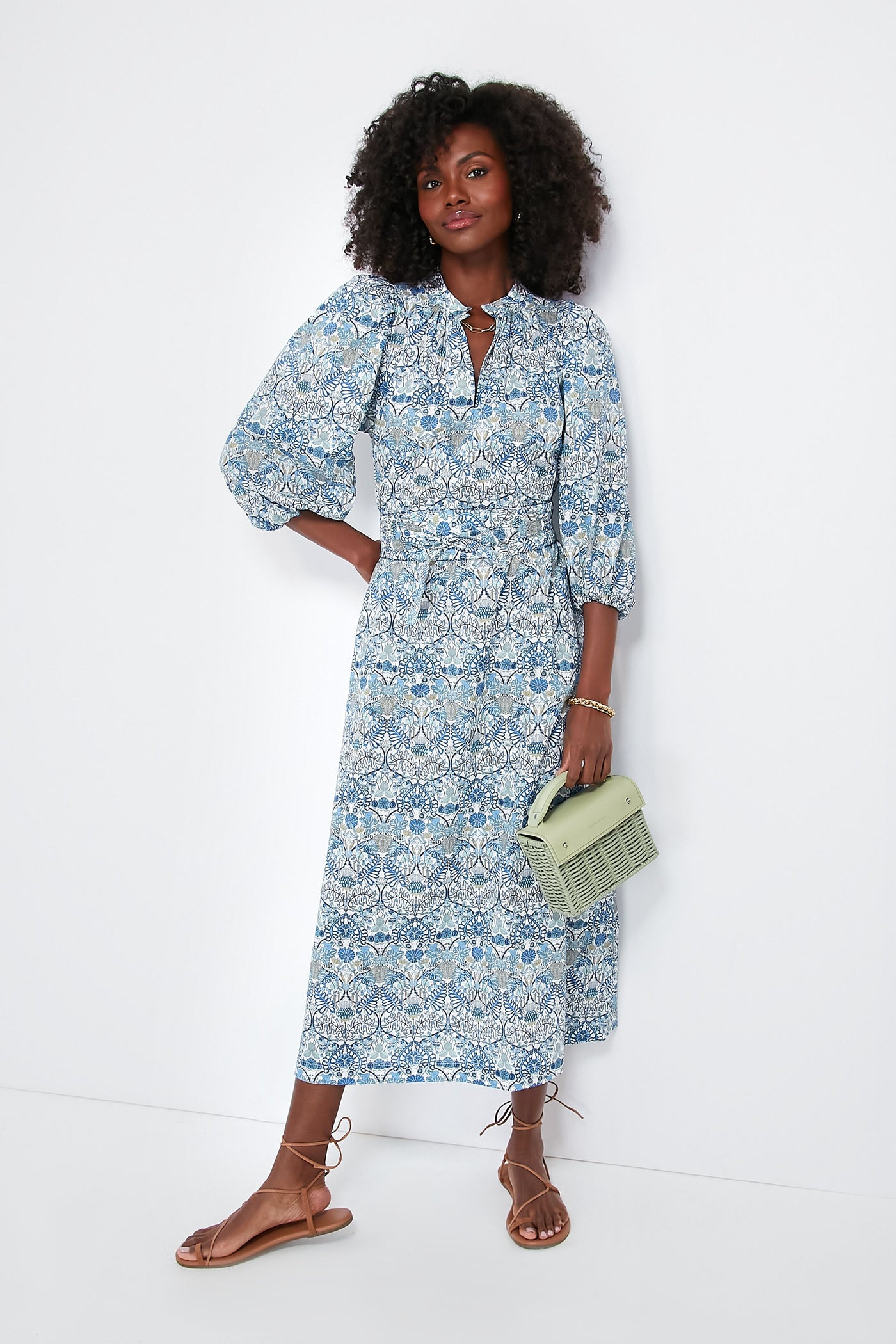 A woman wears an APOF Maya Dress in a blue and white pattern with puff sleeves, pairs it with tan sandals, and carries a light green handbag.
