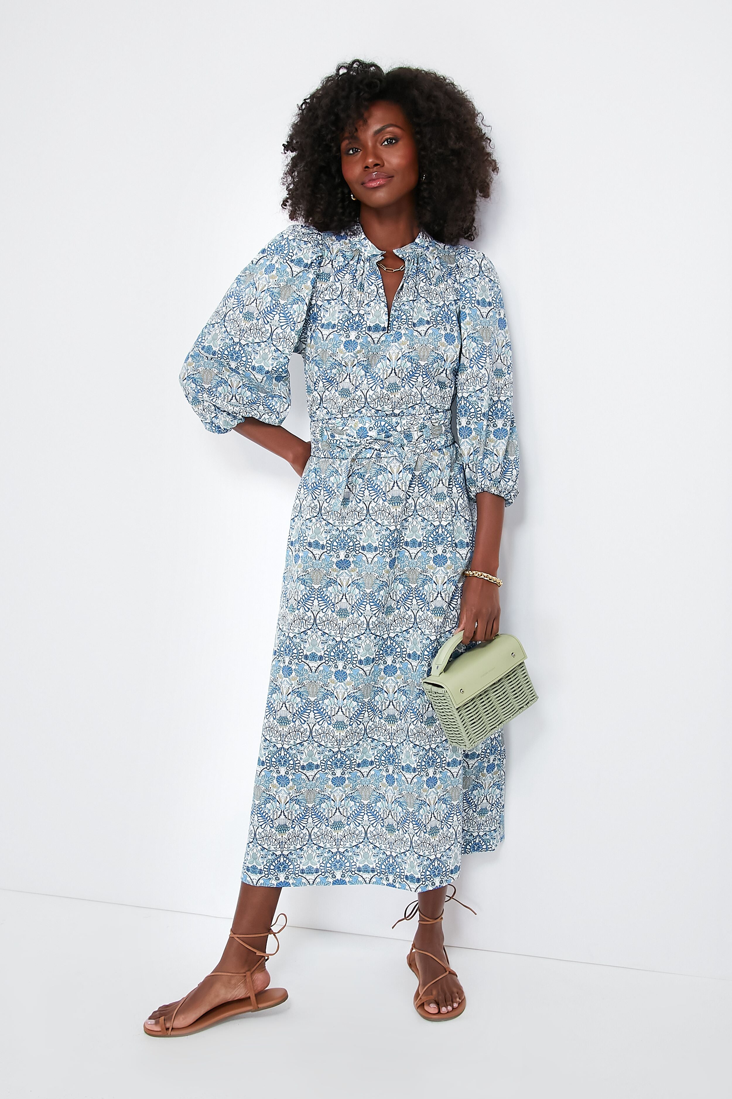 A woman wears an APOF Maya Dress in a blue and white pattern with puff sleeves, pairs it with tan sandals, and carries a light green handbag.