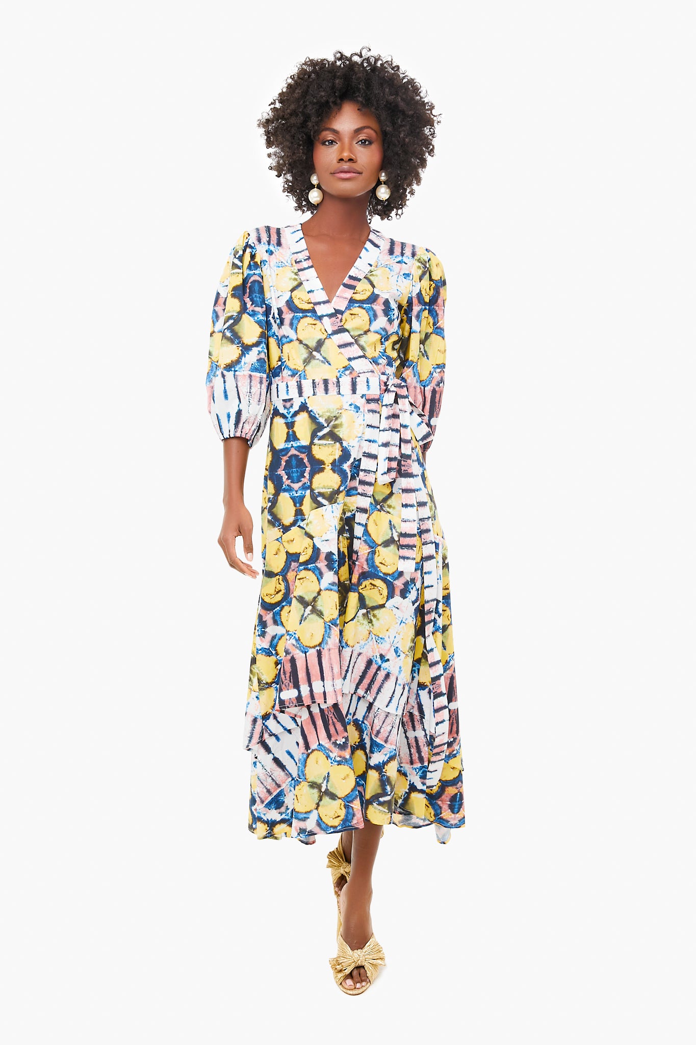 A woman wearing the ALEXIS Marsai Dress—a colorful, patterned piece with puffed sleeves, gold sandals, and an A-line midi skirt—makes this standout style a must-have in womenswear.