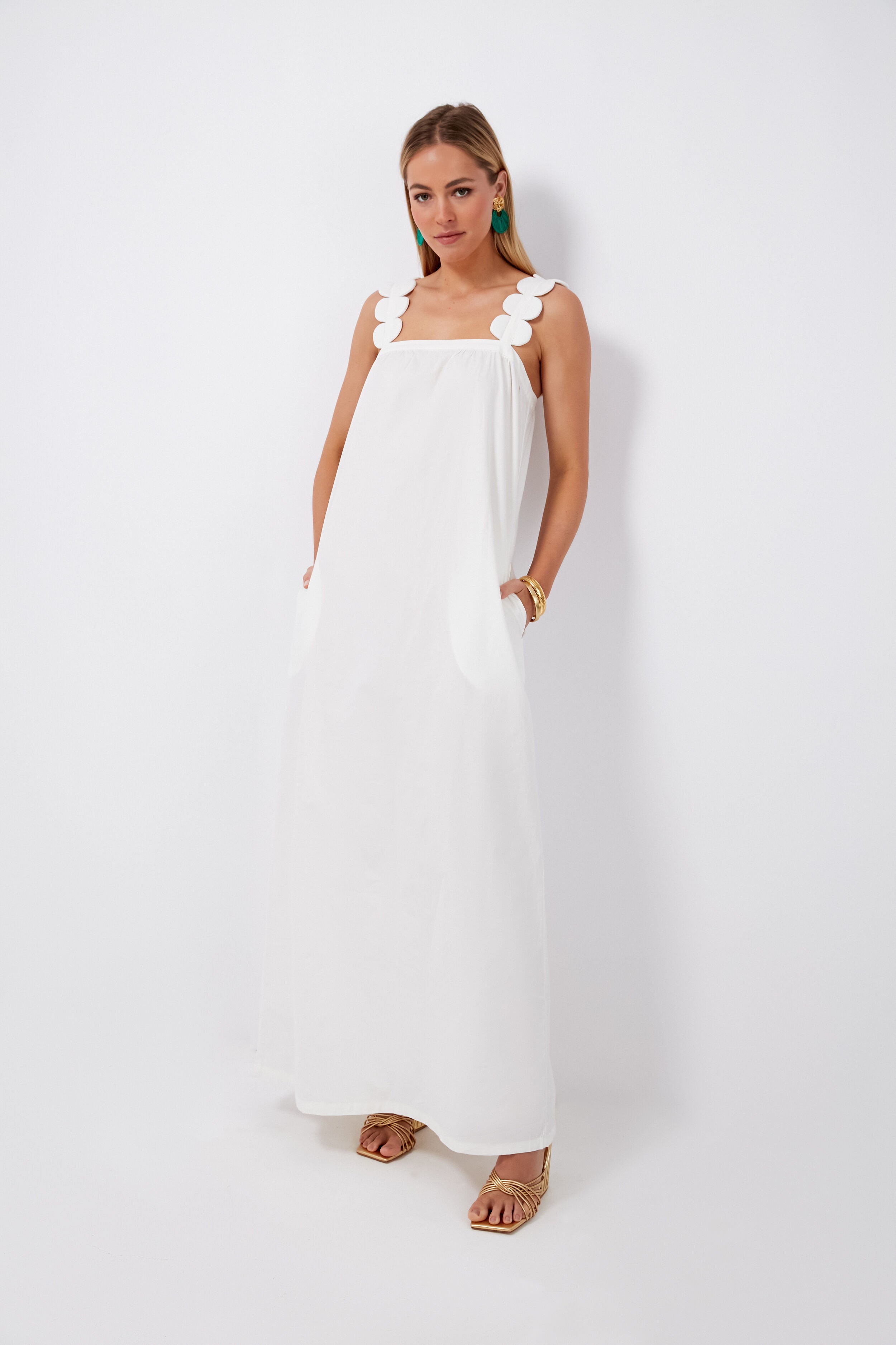 A woman wears an ADRIANA DEGREAS maxi dress with scalloped straps, tan sandals, green earrings, and gold bracelets for an elegant luxury beachwear look.