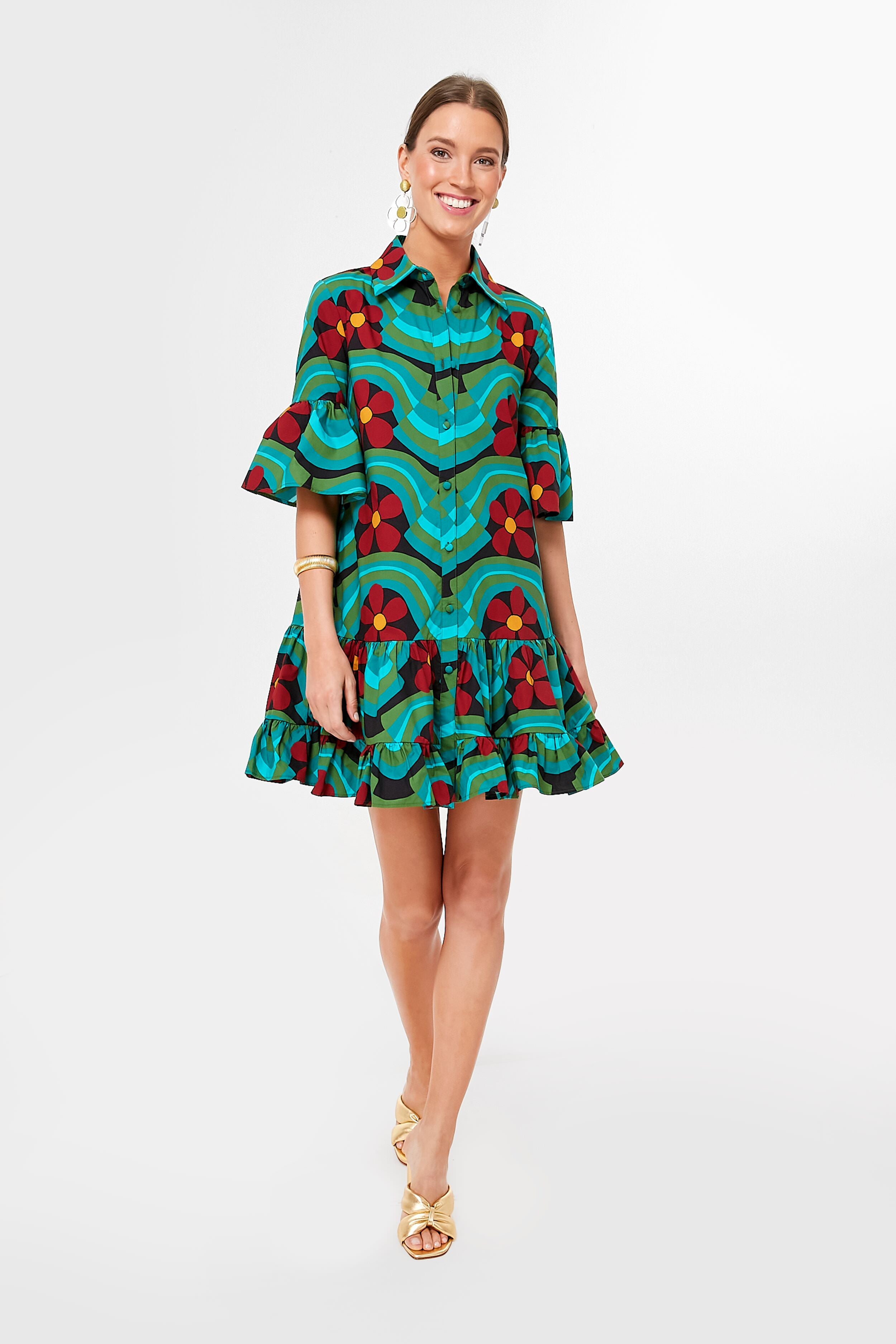 A woman wears a vibrant, patterned La DoubleJ Ashbury Choux Dress in cotton poplin featuring ruffled sleeves and hem, styled with gold sandals and jewelry.