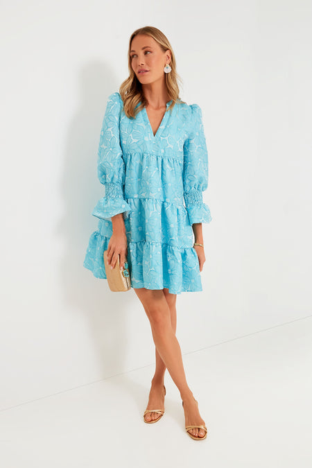 Woman in a light blue daisy jacquard Pomander Place dress with long sleeves and tiered skirt, styled with nude heels, statement earrings, and a beige clutch—making it ideal for transitioning from day to night.
