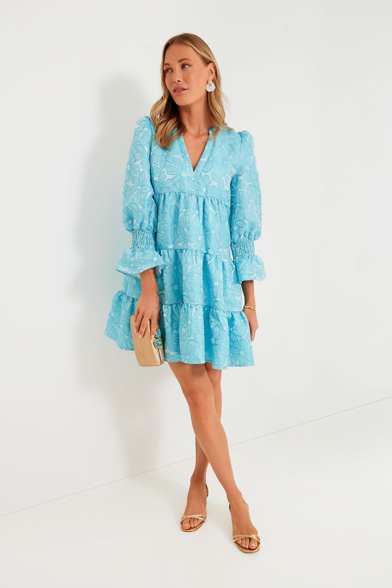 Woman in a light blue daisy jacquard Pomander Place dress with long sleeves and tiered skirt, styled with nude heels, statement earrings, and a beige clutch—making it ideal for transitioning from day to night.