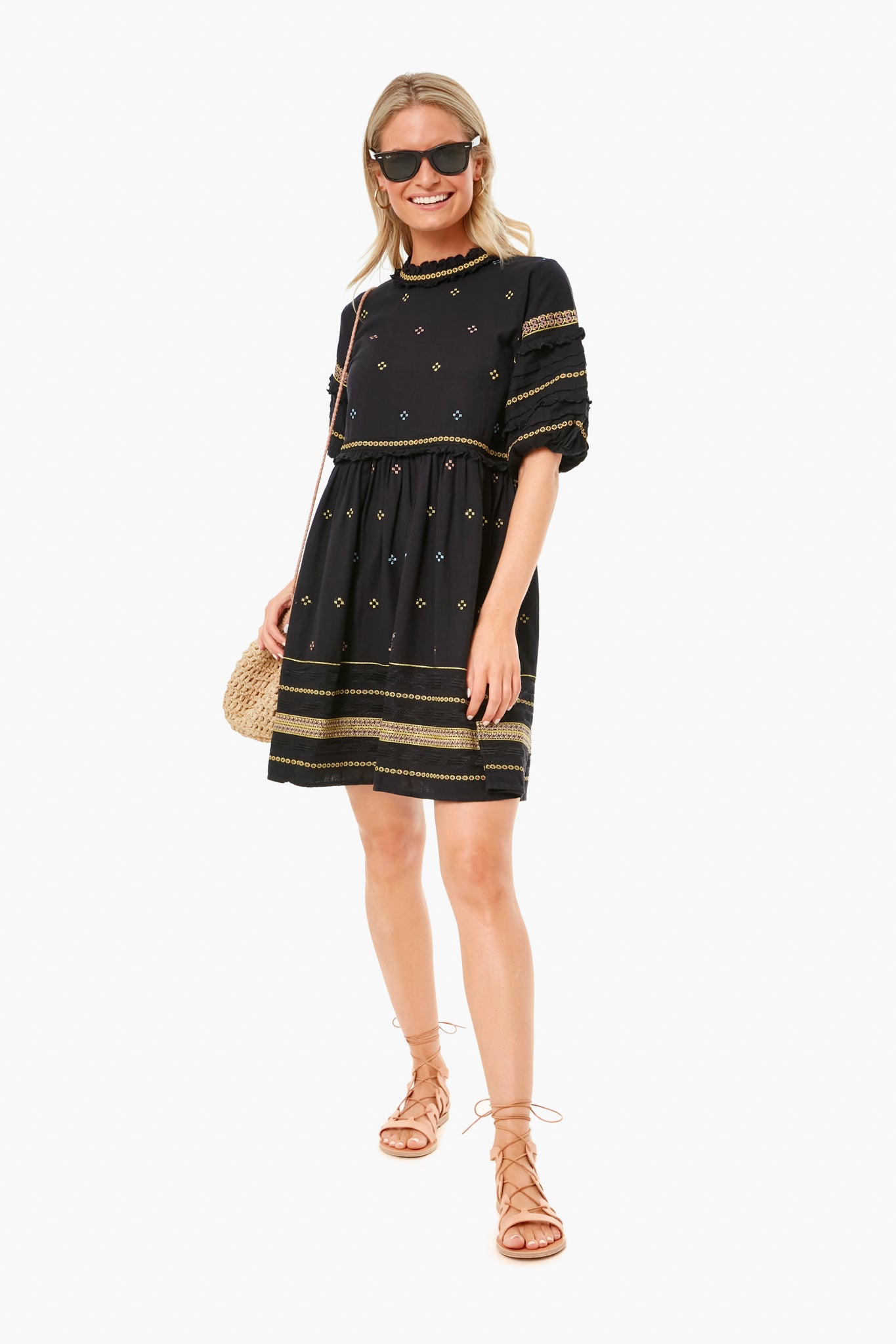 A woman in sunglasses wears THE GREAT. Black Peninsula Dress with gold patterns, lace-up sandals, and carries a woven crossbody bag.